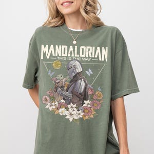 May include: An olive green t-shirt featuring a graphic print of The Mandalorian and Grogu surrounded by colorful flowers, butterflies, and a yellow sun. The text "MANDALORIAN" and "THIS IS THE WAY" is printed in a distressed white font above the characters.