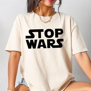 Op de afbeelding: Crèmekleurig T-shirt met de tekst "STOP WARS" in grote, zwarte hoofdletters. Het shirt met korte mouwen heeft een losse pasvorm. De persoon draagt een spijkerbroekshort en witte sneakers.