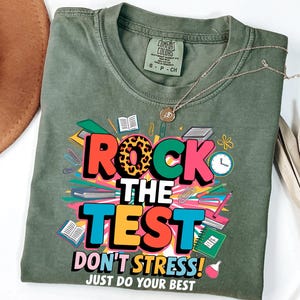 Op de afbeelding: Olijfgroen t-shirt met de tekst "ROCK THE TEST" in kleurrijke blokletters, met een "O" met luipaardprint. Het shirt zegt ook "DON'T STRESS! JUST DO YOUR BEST". Een gouden ketting ligt op het shirt.