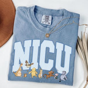 May include: A blue short-sleeve t-shirt with a crew neckline and a Comfort Colors brand label. The front features the text NICU in large white and blue striped block letters, with small illustrations of various cartoon animal characters below.