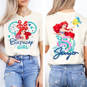 May include: Cream-colored t-shirt featuring a colorful graphic of a cartoon mermaid with red hair, a blue top, and a yellow fish. The front says "Birthday Girl" and the back says "5 Sawyer".