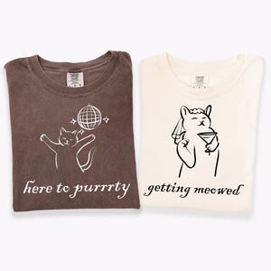 May include: Two folded t-shirts. One is brown with a white cat graphic, a disco ball, and the text "here to purrrty." The other is cream-colored with a cat graphic holding a martini and the text "getting meowed."