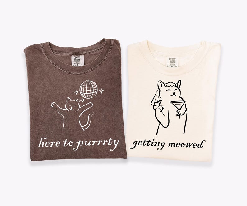 May include: Two folded t-shirts. One is brown with a white cat graphic, a disco ball, and the text "here to purrrty." The other is cream-colored with a cat graphic holding a martini and the text "getting meowed."