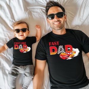 Peut inclure: Deux t-shirts noirs assortis avec le texte "FAST DAD" et un dessin anim&eacute; de voiture de course. L'un est pour un adulte, l'autre pour un enfant. Les deux portent des lunettes de soleil et sont allong&eacute;s sur une surface blanche.