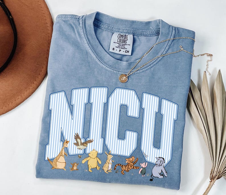 May include: A blue short-sleeve t-shirt with a crew neckline and a Comfort Colors brand label. The front features the text NICU in large white and blue striped block letters, with small illustrations of various cartoon animal characters below.