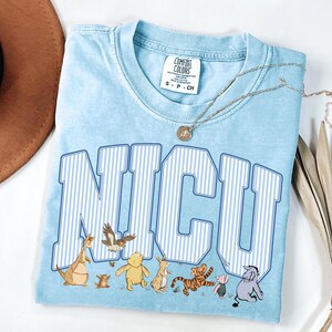 May include: A light blue t-shirt featuring the text NICU in white and blue vertical stripes. Below the text are illustrations of Winnie the Pooh characters including Pooh, Piglet, Tigger, Eeyore, Rabbit, Kanga, Roo, and Owl. The shirt has a Comfort Colors label.