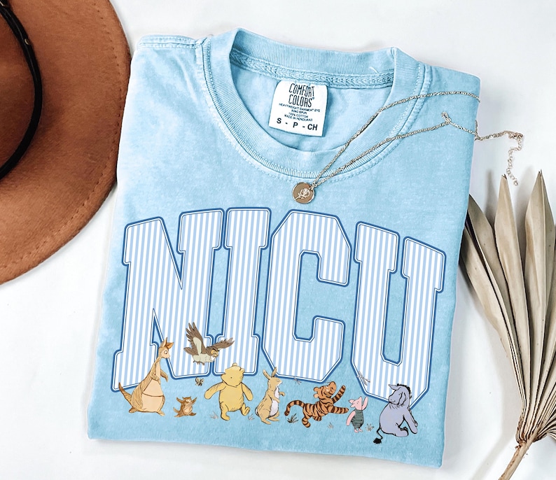 May include: A light blue t-shirt featuring the text NICU in white and blue vertical stripes. Below the text are illustrations of Winnie the Pooh characters including Pooh, Piglet, Tigger, Eeyore, Rabbit, Kanga, Roo, and Owl. The shirt has a Comfort Colors label.