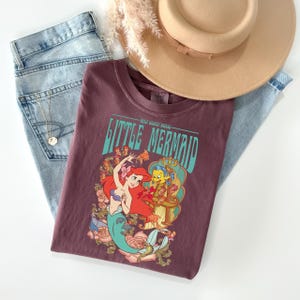 May include: A maroon t-shirt featuring a graphic of Ariel and Flounder from The Little Mermaid, with the text "Little Mermaid". The shirt is paired with light wash jeans and a tan hat.
