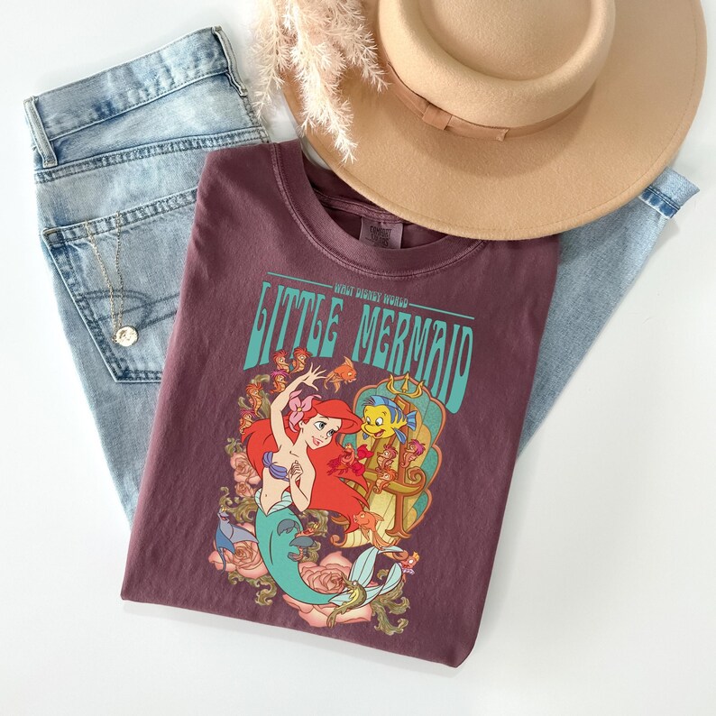 May include: A maroon t-shirt featuring a graphic of Ariel and Flounder from The Little Mermaid, with the text "Little Mermaid". The shirt is paired with light wash jeans and a tan hat.