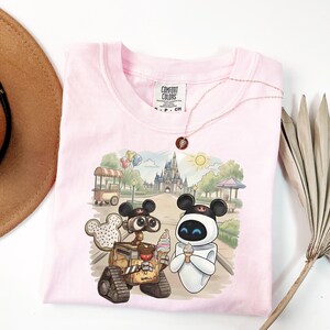 Puede incluir: Camiseta Comfort Colors rosa claro con un gr&aacute;fico de Wall-E y Eva de Disney, sosteniendo conos de helado frente a un castillo. La camiseta tiene una etiqueta Comfort Colors y un collar.