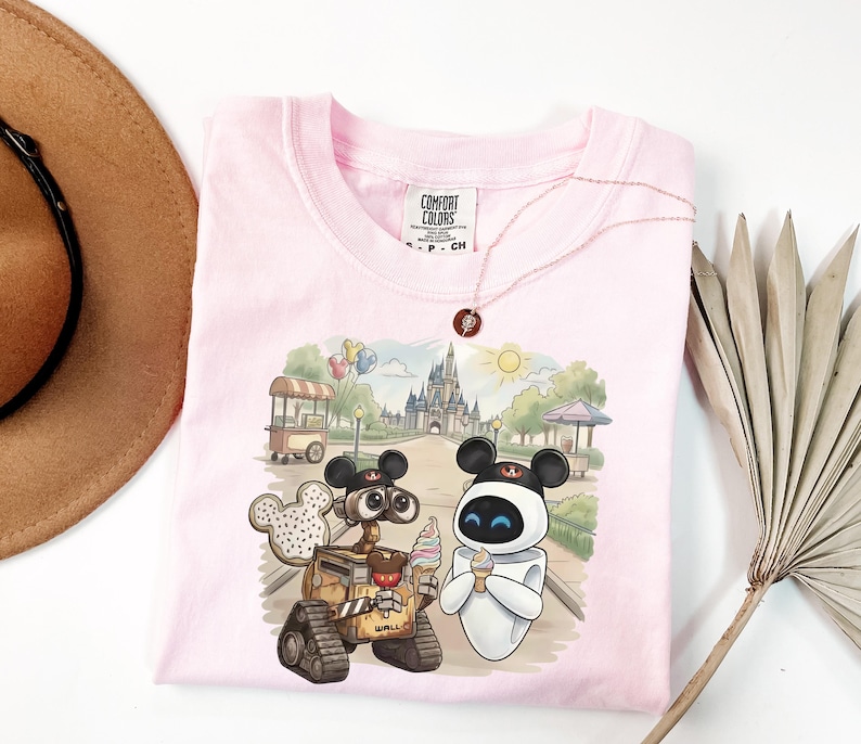 Puede incluir: Camiseta Comfort Colors rosa claro con un gr&aacute;fico de Wall-E y Eva de Disney, sosteniendo conos de helado frente a un castillo. La camiseta tiene una etiqueta Comfort Colors y un collar.