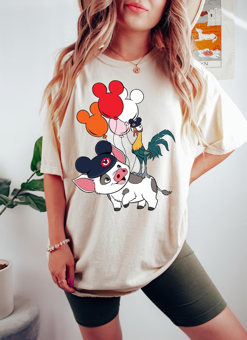 Pode incluir: Camiseta creme com uma ilustra&ccedil;&atilde;o de desenho animado de um porco usando um chap&eacute;u do Mickey Mouse, um galo e uma vaca. O porco est&aacute; montando a vaca, e o galo est&aacute; nas costas da vaca. Bal&otilde;es vermelhos, laranja, rosa e brancos flutuam.