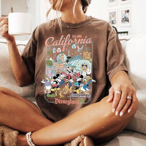 Puede incluir: Camiseta marr&oacute;n con un dise&ntilde;o colorido de Disneyland, con personajes y atracciones. El texto "California Disneyland Est. 1955" est&aacute; presente. La persona est&aacute; sentada en un sof&aacute; beige.