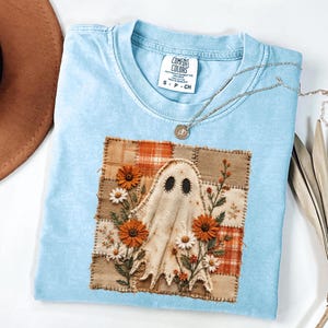 May include: A light blue t-shirt featuring a square patch design with a central ghost figure surrounded by embroidered orange and white flowers. The patch has a patchwork background of tan and orange plaid squares. The shirt label reads Comfort Colors.