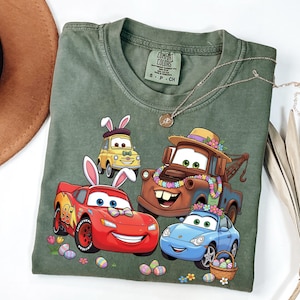 Può includere: T-shirt verde oliva con grafica di auto animate decorate con orecchie da coniglietto pasquale e dettagli floreali. Il design include Saetta McQueen, Cricchetto e altri personaggi, insieme a uova di Pasqua e un cestino.