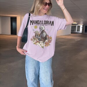May include: A light purple short-sleeve t-shirt featuring a graphic print of The Mandalorian and Grogu surrounded by colorful flowers. The text "MANDALORIAN" and "THIS IS THE WAY" is printed above the characters in a dark, distressed font style.