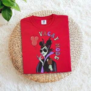 Puede incluir: Camiseta roja de cuello redondo con un gr&aacute;fico de un villano de dibujos animados con gafas de sol y sosteniendo un dulce de chocolate. El texto "VACAY MODE" est&aacute; impreso en la parte delantera, junto con un globo de Mickey Mouse.