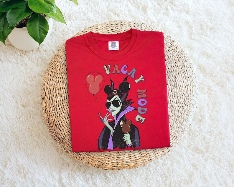 Puede incluir: Camiseta roja de cuello redondo con un gr&aacute;fico de un villano de dibujos animados con gafas de sol y sosteniendo un dulce de chocolate. El texto "VACAY MODE" est&aacute; impreso en la parte delantera, junto con un globo de Mickey Mouse.