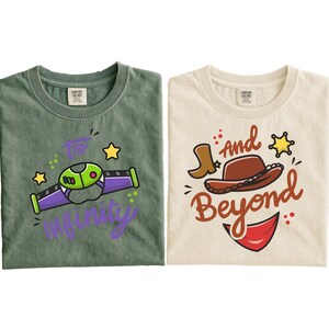 May include: Two t-shirts, one green and one cream-colored, with cartoon graphics. The green shirt features a purple and green spaceship design with the words "To Infinity" in purple. The cream shirt has a cowboy hat, boot, and the words "And Beyond".