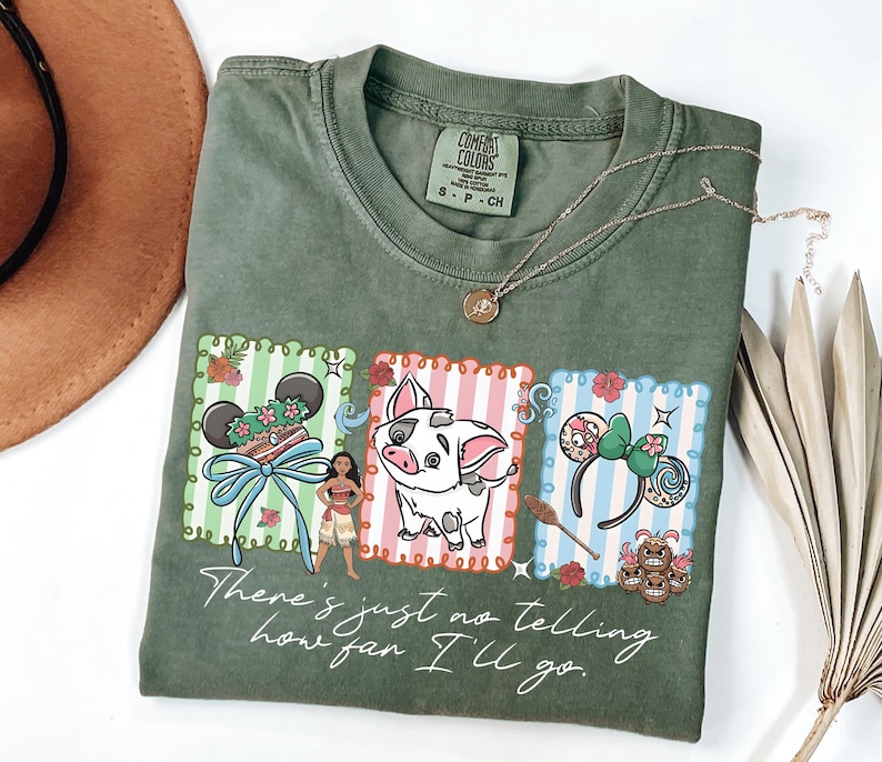 May include: An olive green t-shirt featuring a graphic print of Moana, Pua the pig, and two pairs of character-themed mouse ears. The text "There's just no telling how far I'll go" is printed in white script below the colorful rectangular illustrations.