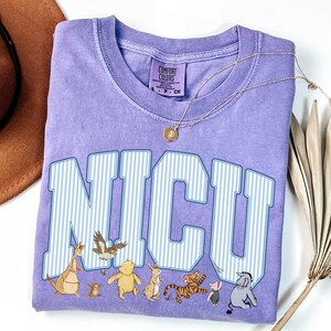May include: A lavender short-sleeved t-shirt featuring the text NICU in large white and blue striped varsity-style letters. Below the text are illustrations of various cartoon characters including a bear, a tiger, a donkey, and a piglet.