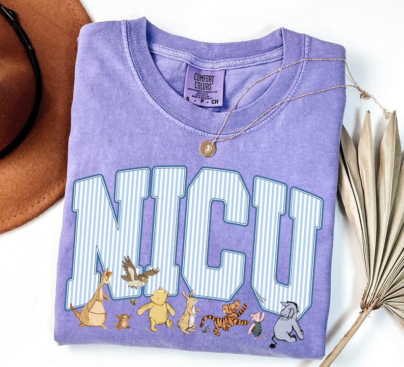 May include: A lavender short-sleeved t-shirt featuring the text NICU in large white and blue striped varsity-style letters. Below the text are illustrations of various cartoon characters including a bear, a tiger, a donkey, and a piglet.
