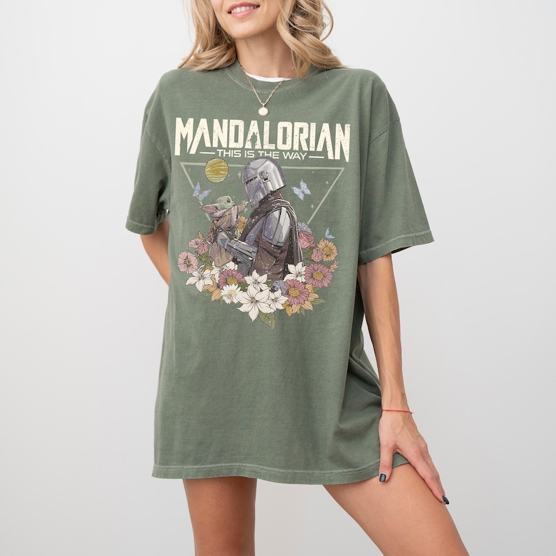 May include: An olive green t-shirt featuring a graphic print of The Mandalorian and Grogu surrounded by colorful flowers, butterflies, and a yellow sun. The text "MANDALORIAN" and "THIS IS THE WAY" is printed in a distressed white font above the characters.