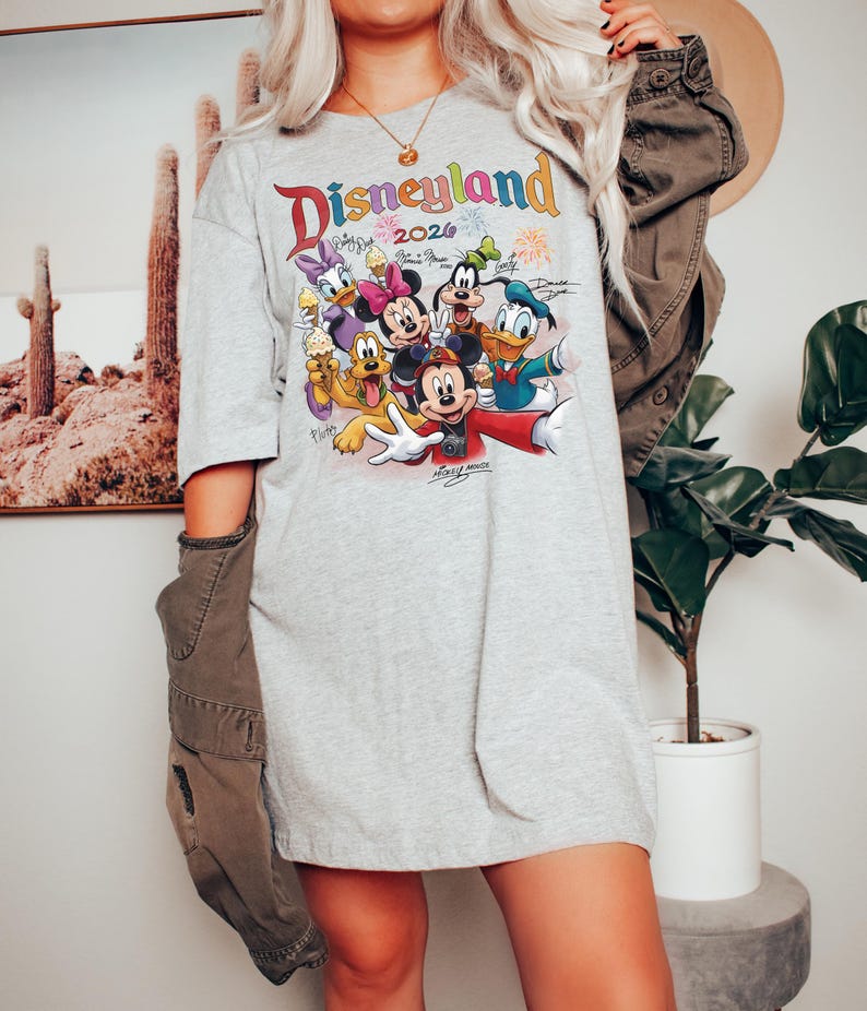 K&ouml;nnte beinhalten: Ein graues T-Shirt mit einem farbenfrohen Aufdruck von Disney-Figuren, darunter Mickey Mouse, Minnie Mouse, Donald Duck und Goofy. Das Shirt tr&auml;gt den Text "Disneyland 2026" in einer Regenbogen-Schriftart. Das Design beinhaltet Unterschriften und Feuerwerk.