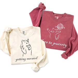 May include: Two long-sleeved shirts. One is cream-colored with a black graphic of a llama holding a martini glass, with the text "getting meowed". The other is maroon with a white cat graphic and the text "here to purrrty".