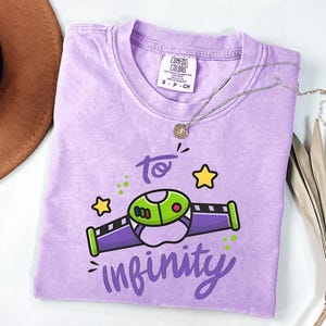May include: A lavender Comfort Colors t-shirt featuring a cartoon graphic of a space ship with the text "to infinity". The graphic is green, purple, and black with yellow stars. A silver necklace with a pendant is on the shirt.