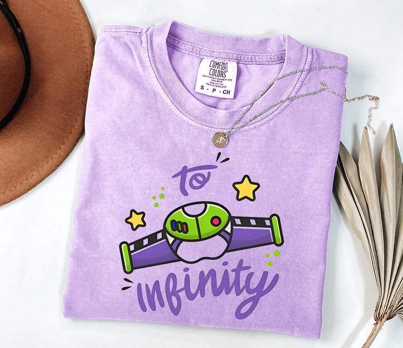 May include: A lavender Comfort Colors t-shirt featuring a cartoon graphic of a space ship with the text "to infinity". The graphic is green, purple, and black with yellow stars. A silver necklace with a pendant is on the shirt.