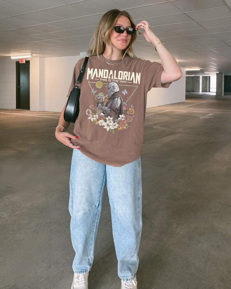 May include: A brown short-sleeve t-shirt featuring a graphic print of The Mandalorian and Grogu surrounded by colorful flowers and butterflies. The text "MANDALORIAN" and "THIS IS THE WAY" is printed above the characters. Light blue acid-wash jeans.