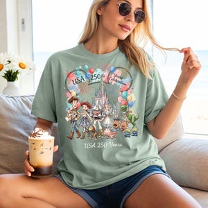 Peut inclure: Un t-shirt vert sauge avec un graphique color&eacute; repr&eacute;sentant des personnages de Toy Story, un ch&acirc;teau, des feux d'artifice et des ballons. Le texte indique USA 250 Years Celebration et USA 250 Years. Le design inclut Woody, Jessie et Buzz.