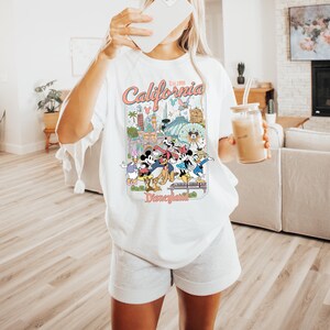 Puede incluir: Camiseta blanca con un gr&aacute;fico colorido de Disneyland, con personajes y lugares emblem&aacute;ticos. El dise&ntilde;o incluye el texto "California Est. 1955" y "Disneyland". La camiseta es de estilo casual y holgado.