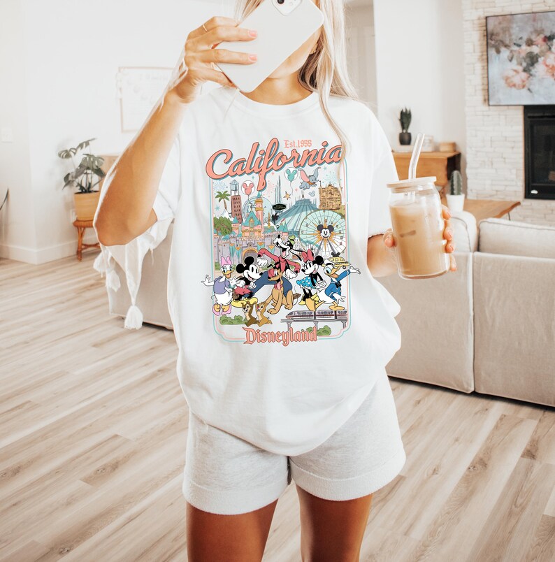 Puede incluir: Camiseta blanca con un gr&aacute;fico colorido de Disneyland, con personajes y lugares emblem&aacute;ticos. El dise&ntilde;o incluye el texto "California Est. 1955" y "Disneyland". La camiseta es de estilo casual y holgado.