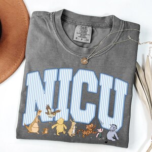 May include: A charcoal gray short-sleeve t-shirt featuring the text NICU in large white and blue striped varsity-style letters. Below the text are small illustrations of Winnie the Pooh characters including Pooh, Piglet, Tigger, Eeyore, Rabbit, and Roo.