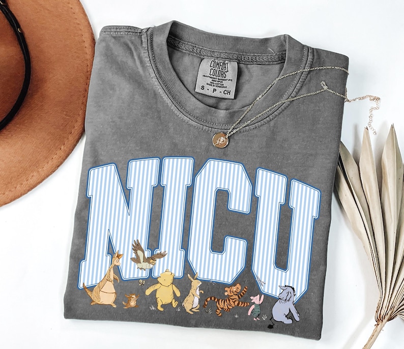 May include: A charcoal gray short-sleeve t-shirt featuring the text NICU in large white and blue striped varsity-style letters. Below the text are small illustrations of Winnie the Pooh characters including Pooh, Piglet, Tigger, Eeyore, Rabbit, and Roo.