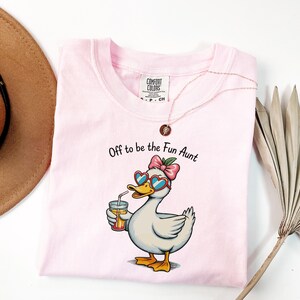 May include: A light pink t-shirt featuring a graphic of a white duck wearing heart-shaped sunglasses and a pink bow, holding a glass with a straw. The text above the duck reads, "Off to be the Fun Aunt." The shirt has a Comfort Colors brand label.