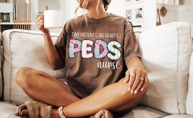 May include: A brown t-shirt with the text "Tiny Patients, Big Hearts" above the word "PEDS" in a floral and striped design, and the word "nurse" below. The shirt is worn by a person sitting on a couch.