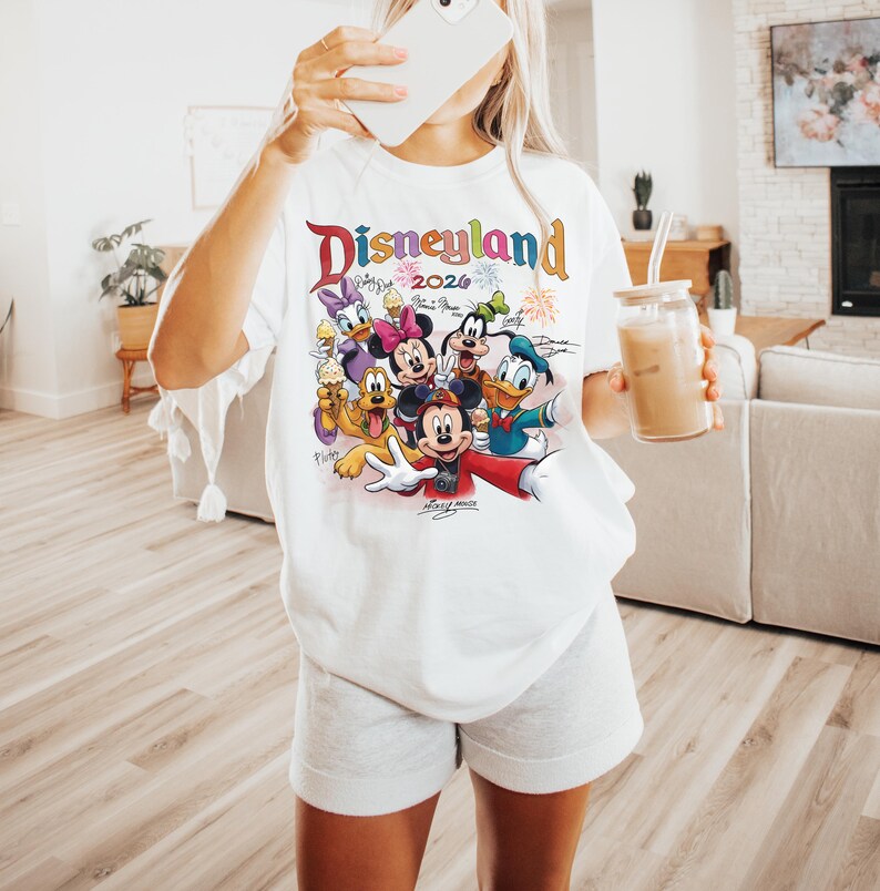 K&ouml;nnte beinhalten: Wei&szlig;es T-Shirt mit einem farbenfrohen Disneyland-Motiv. Das Design enth&auml;lt den Text "Disneyland 2026" und zeigt Disney-Figuren wie Mickey Mouse, Minnie Mouse, Donald Duck, Goofy und Daisy Duck.