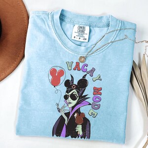 Puede incluir: Camiseta azul claro con un gr&aacute;fico de un personaje de dibujos animados con gafas de sol y un helado con forma de Mickey Mouse. El texto "VACAY MODE" est&aacute; encima del personaje. Tambi&eacute;n se ven un collar plateado y un sombrero marr&oacute;n.