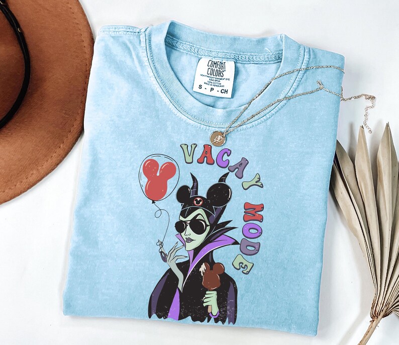 Puede incluir: Camiseta azul claro con un gr&aacute;fico de un personaje de dibujos animados con gafas de sol y un helado con forma de Mickey Mouse. El texto "VACAY MODE" est&aacute; encima del personaje. Tambi&eacute;n se ven un collar plateado y un sombrero marr&oacute;n.
