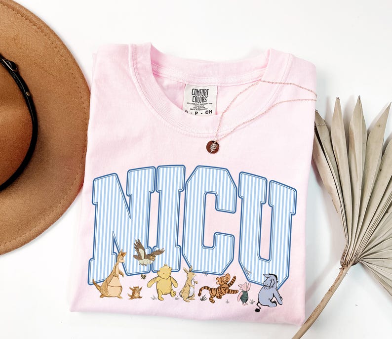 May include: A light pink t-shirt featuring the text NICU in large blue and white striped varsity-style letters. Below the text are small illustrations of various cartoon characters including a bear, a tiger, a donkey, and a small piglet.