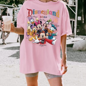 K&ouml;nnte beinhalten: Ein hellrosa T-Shirt mit dem Wort "Disneyland" in bunten Buchstaben und dem Jahr 2026. Das Shirt zeigt eine Grafik von Disney-Charakteren, darunter Mickey Mouse, Minnie Mouse, Donald Duck und Goofy.