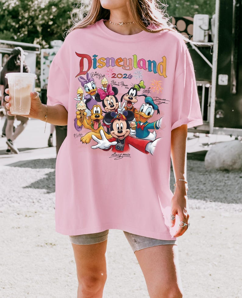 K&ouml;nnte beinhalten: Ein hellrosa T-Shirt mit dem Wort "Disneyland" in bunten Buchstaben und dem Jahr 2026. Das Shirt zeigt eine Grafik von Disney-Charakteren, darunter Mickey Mouse, Minnie Mouse, Donald Duck und Goofy.