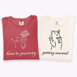 May include: Two folded t-shirts. One is red with a white cat graphic, a disco ball, and the text "here to purrrty." The other is cream-colored with a cat graphic holding a martini and the text "getting meowed."