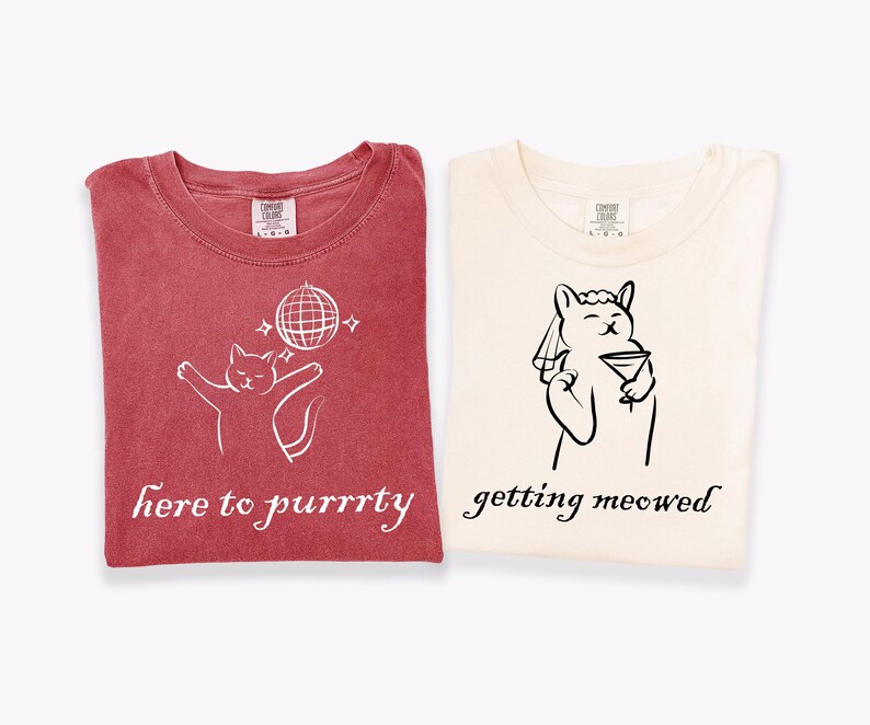 May include: Two folded t-shirts. One is red with a white cat graphic, a disco ball, and the text "here to purrrty." The other is cream-colored with a cat graphic holding a martini and the text "getting meowed."