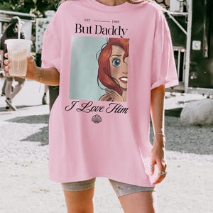 Puede incluir: Camiseta rosa oversize con el texto "But Daddy I Love Him" y un gr&aacute;fico de dibujos animados de un personaje con pelo rojo y ojos azules. La camiseta tambi&eacute;n tiene el texto "EST 1989".