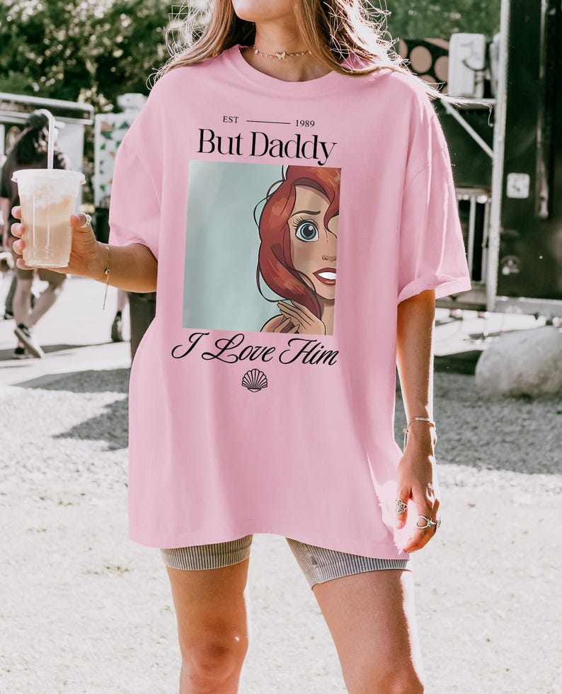 Puede incluir: Camiseta rosa oversize con el texto "But Daddy I Love Him" y un gr&aacute;fico de dibujos animados de un personaje con pelo rojo y ojos azules. La camiseta tambi&eacute;n tiene el texto "EST 1989".