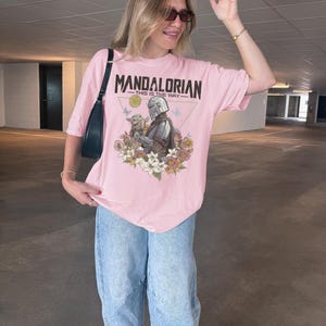 May include: A light pink short-sleeved t-shirt featuring a graphic print of The Mandalorian and Grogu surrounded by colorful flowers and butterflies. The text reads MANDALORIAN THIS IS THE WAY in black lettering above the central character illustration.